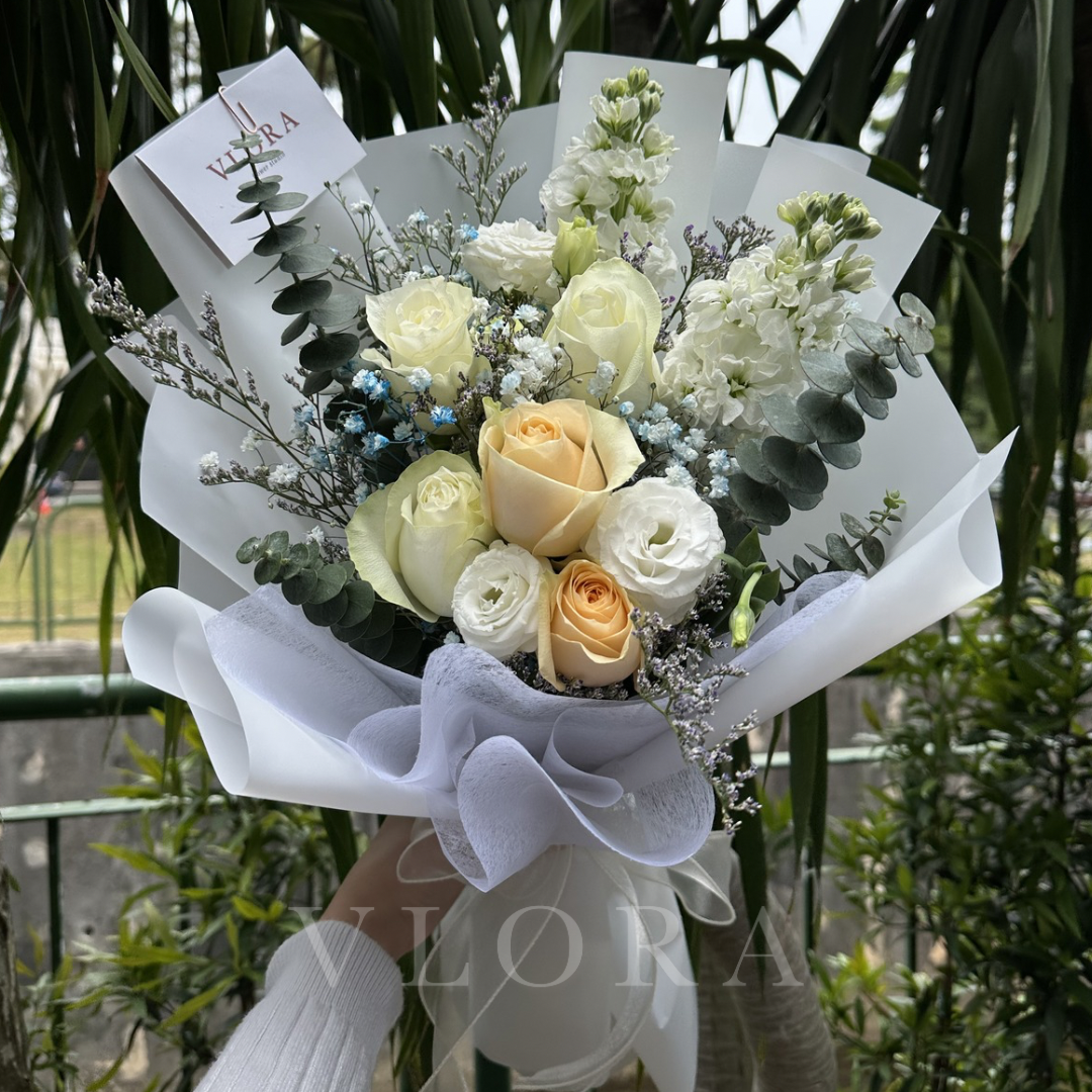 Bouquet of white and yellow roses with greenery, held by a person wearing gloves, with a blurred background.
