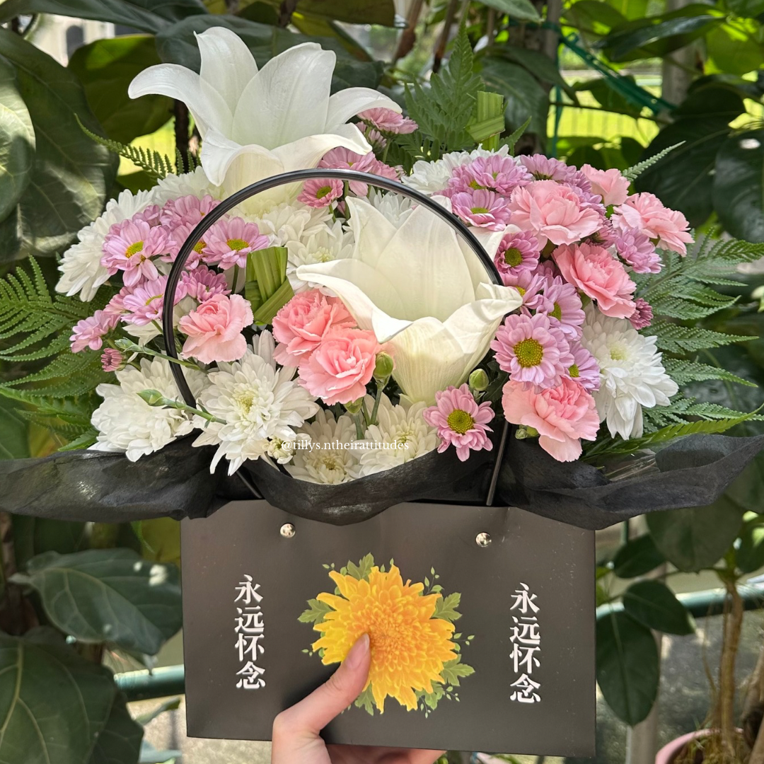 A floral bag arrangement with white lilies, pink carnations, and chrysanthemums, presented in a condolence bag with a printed message.