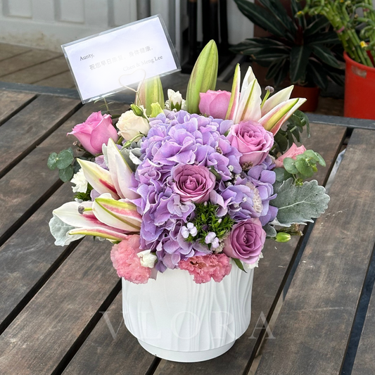 Floral arrangement in a white vase on a wooden table with a plant in the background.