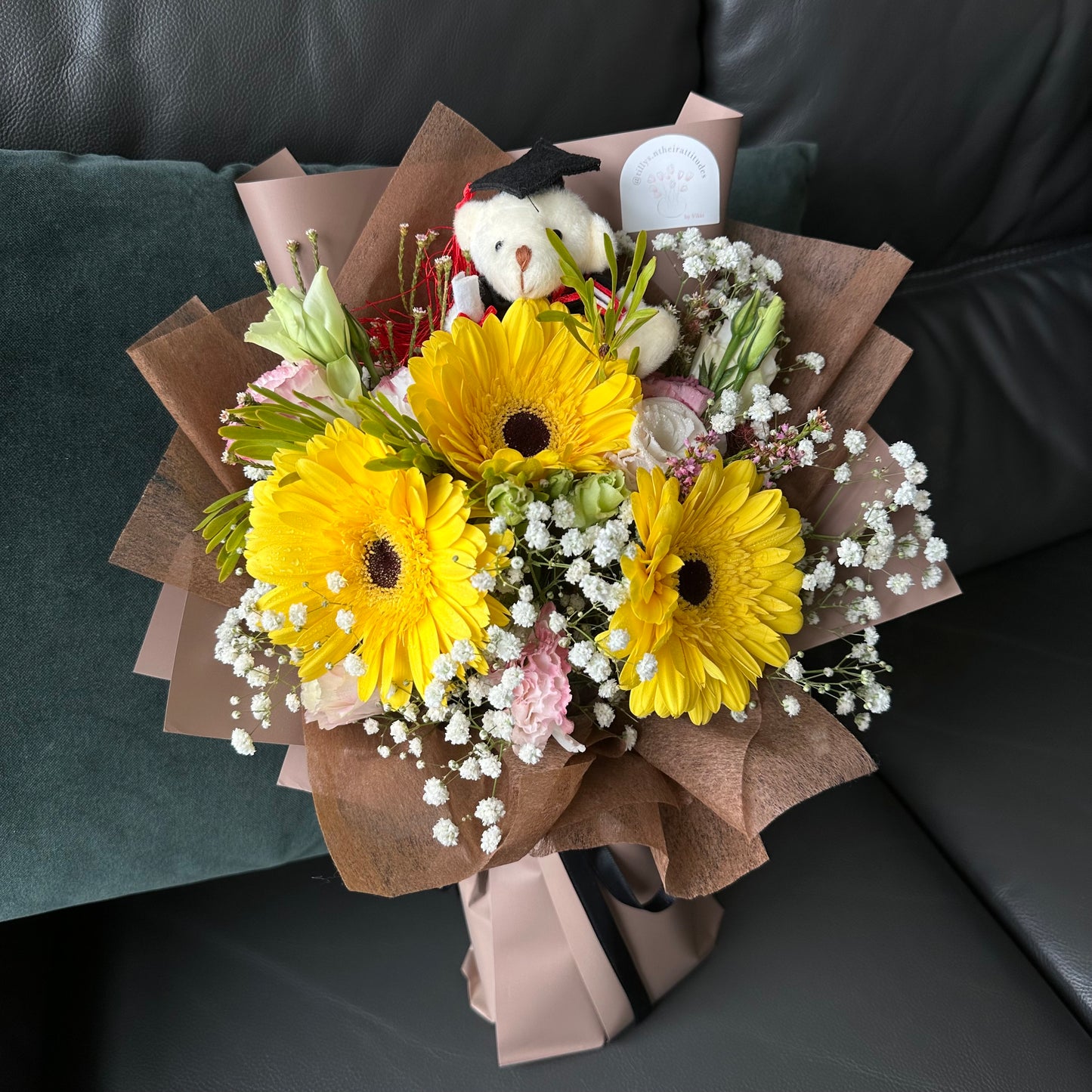 A bouquet of flowers featuring sunny yellow Gerberas, elegant Eustoma, and Baby’s Breath, wrapped in brown paper, accompanied by a cuddly Graduation Bear.