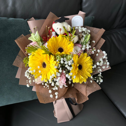 A bouquet of flowers featuring sunny yellow Gerberas, elegant Eustoma, and Baby’s Breath, wrapped in brown paper, accompanied by a cuddly Graduation Bear.