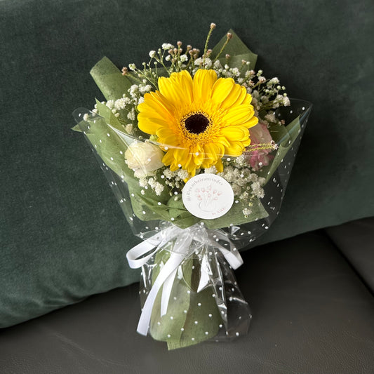A wrapped bouquet consisting of a yellow Gerbera, Eustoma, and Baby’s Breath flowers with a white ribbon, displayed on a green surface.
