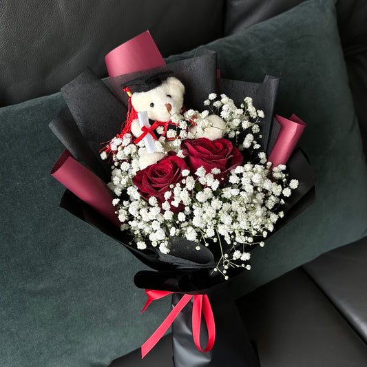 A bouquet arrangement consisting of red roses and Baby's Breath with a white teddy bear, wrapped with red and black ribbons.