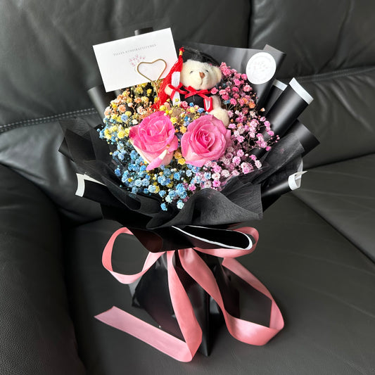 A bouquet of flowers with two bold hot pink roses, vibrant rainbow Baby's Breath, and a charm Graduation Bear, elegantly wrapped in black with a pink ribbon and a complimentary message card.