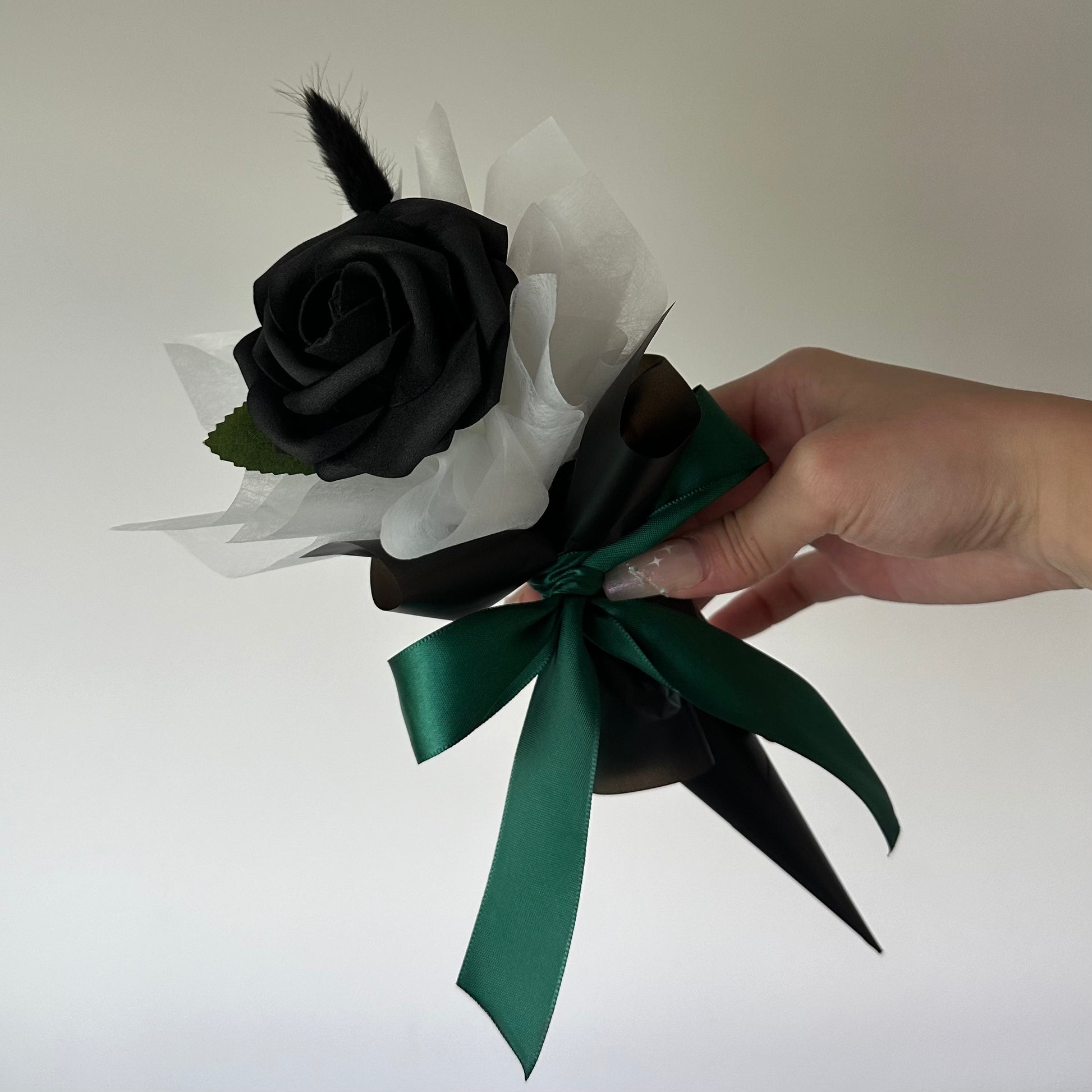 A hand holding a bouquet with a single black foam rose, white and black wrapping, and a green ribbon.