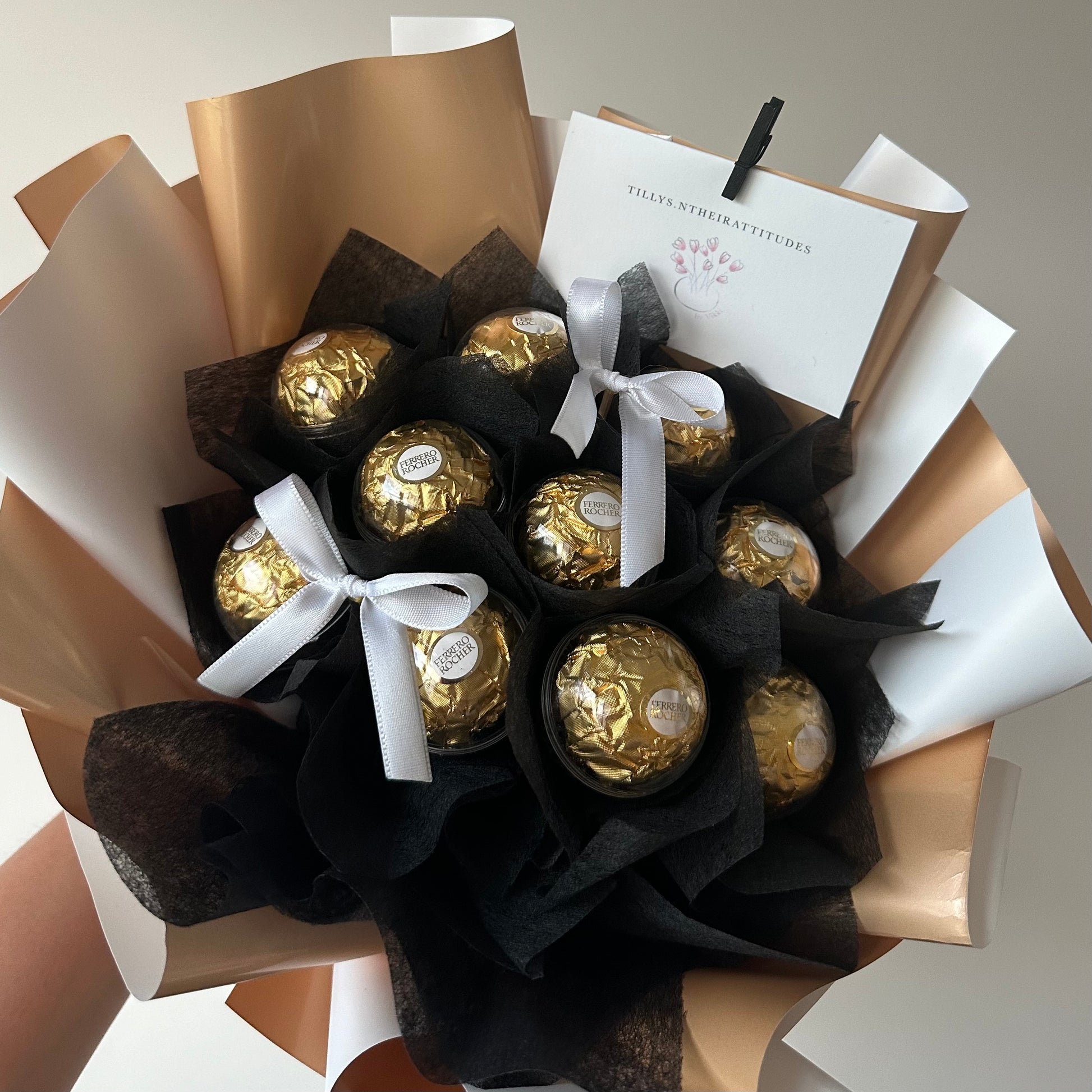 A chocolate bouquet arrangement with Ferrero Rocher candies wrapped in black and white cotton flowers, adorned with silver ribbons, and including a message card.
