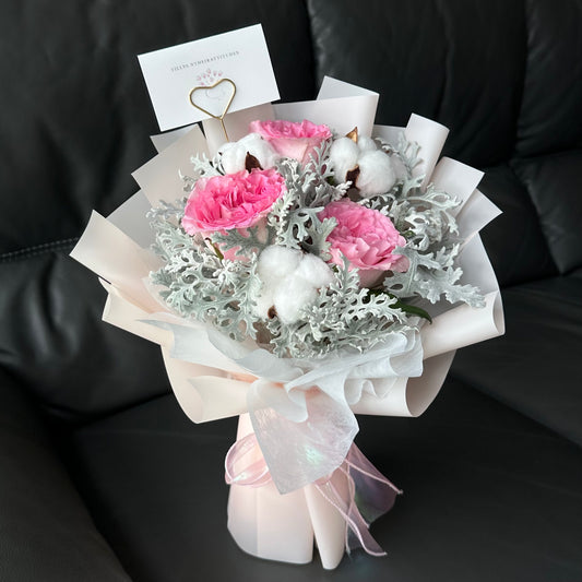 A bouquet of pink roses and white cotton flowers, wrapped in light pink paper with a message card attached.