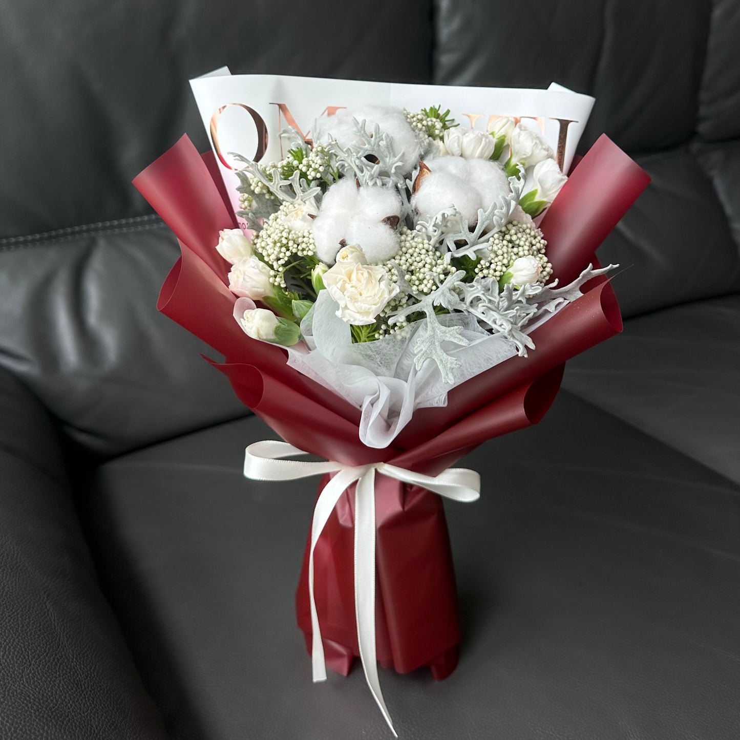 A bouquet arrangement of white cotton flowers and white carnations, wrapped in red and white paper with a decorative ribbon, presented on a dark couch.