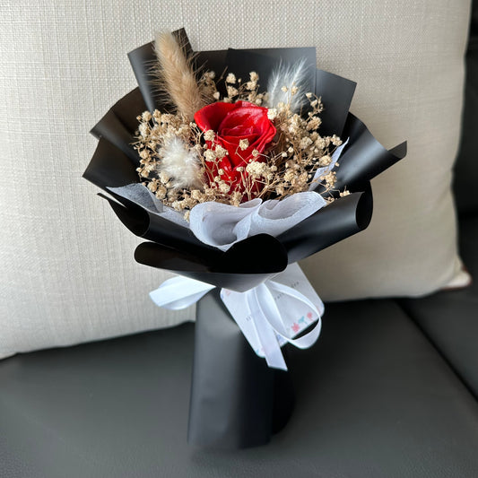 A preserved red rose bouquet elegantly wrapped in black with decorative elements and a white ribbon, placed on a sofa cushion.