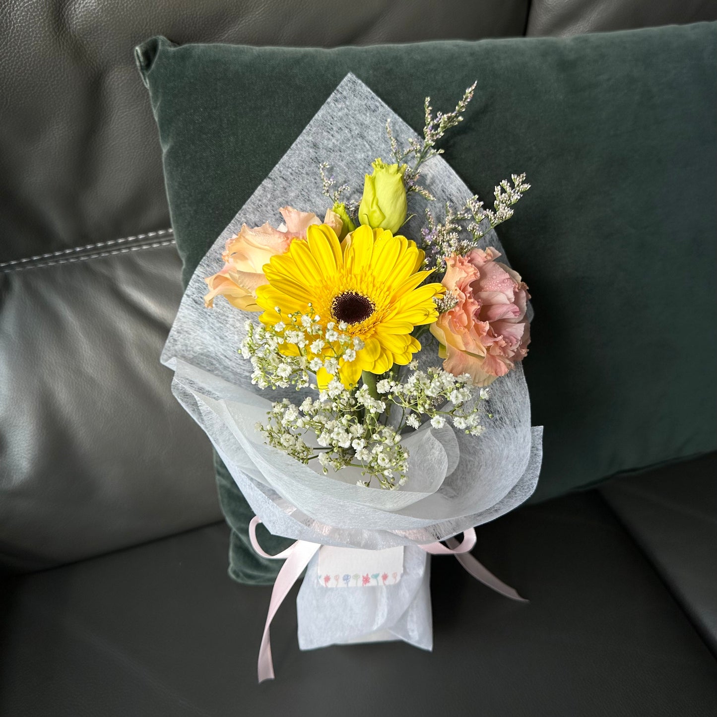 A bright yellow Gerbera daisy paired with pink Eustomas roses, wrapped in white paper with decorative elements, presented on a green cushion.