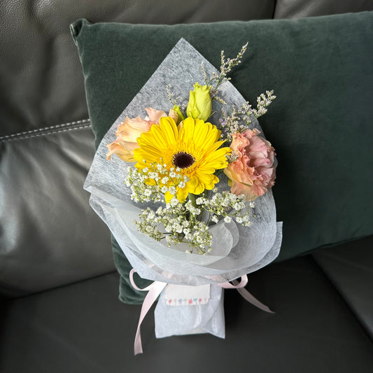 A bright yellow Gerbera daisy paired with pink Eustomas roses, wrapped in white paper with decorative elements, presented on a green cushion.