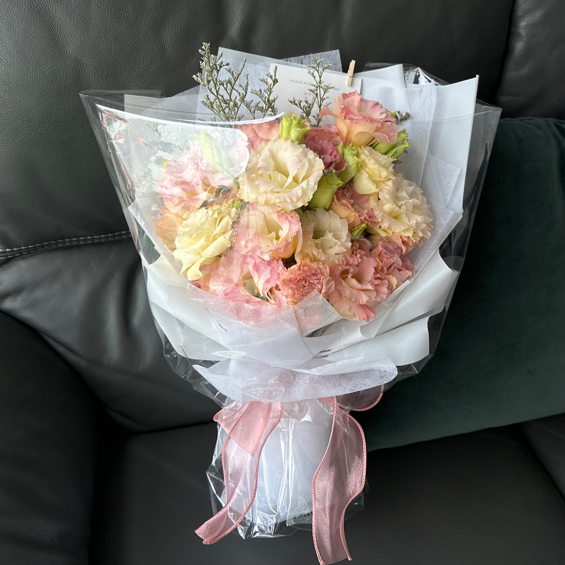 A bouquet of pastel pink and yellow Eustomas wrapped in white paper with a pink ribbon, placed on a sofa.