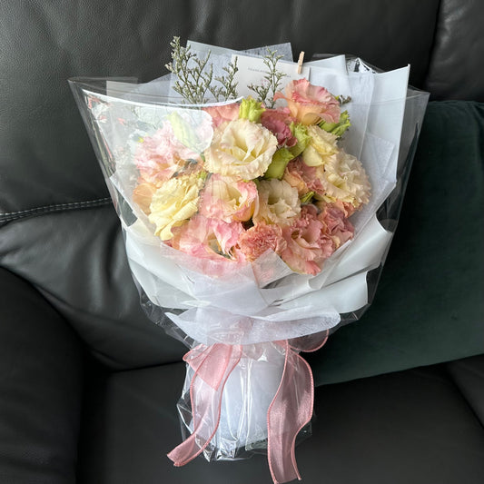 A bouquet of pastel pink and yellow Eustomas wrapped in white paper with a pink ribbon, placed on a sofa.
