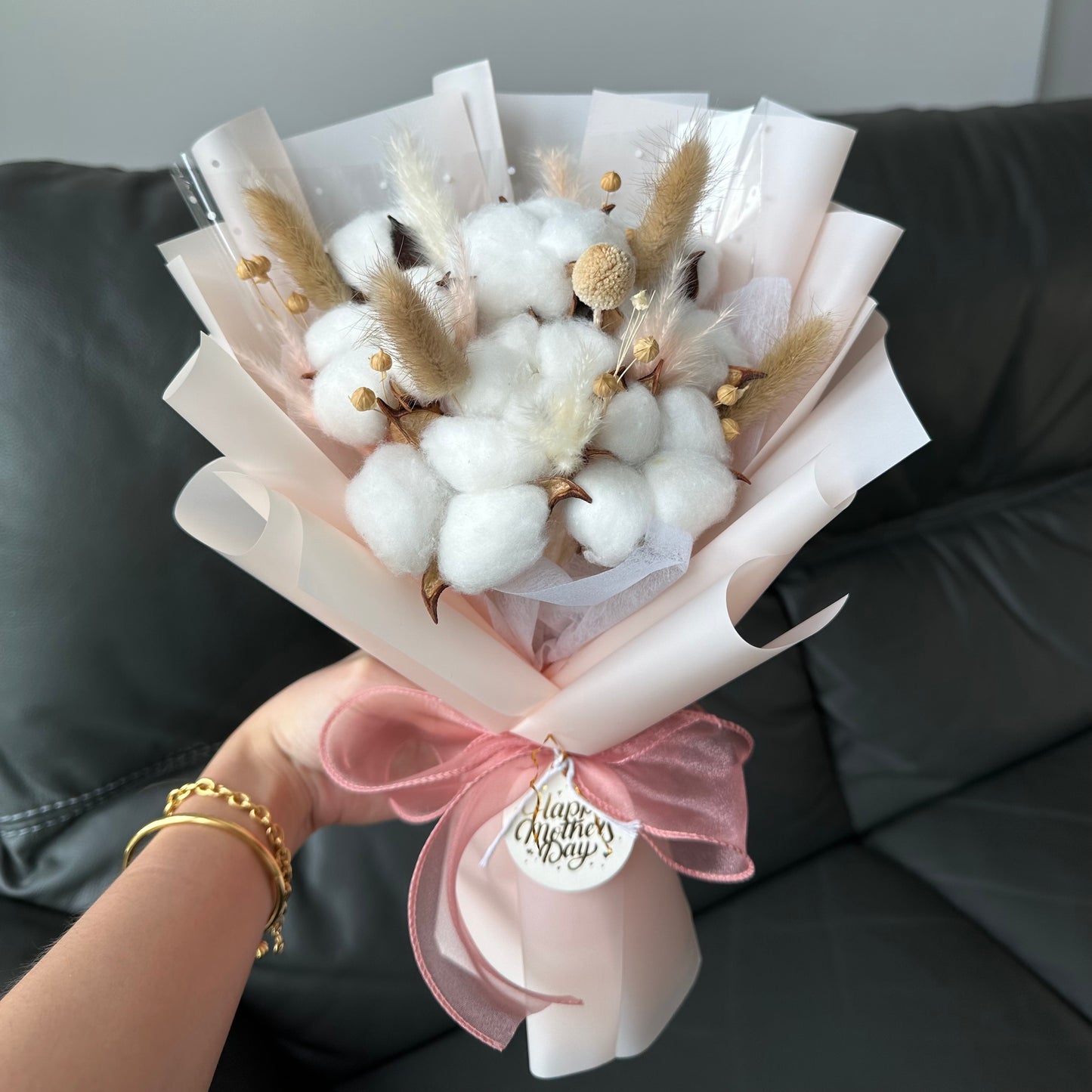 A preserved hand bouquet with white cotton flowers and bunny tails, wrapped in a light color with a pink ribbon and a tag that says 'Happy Mother's Day.