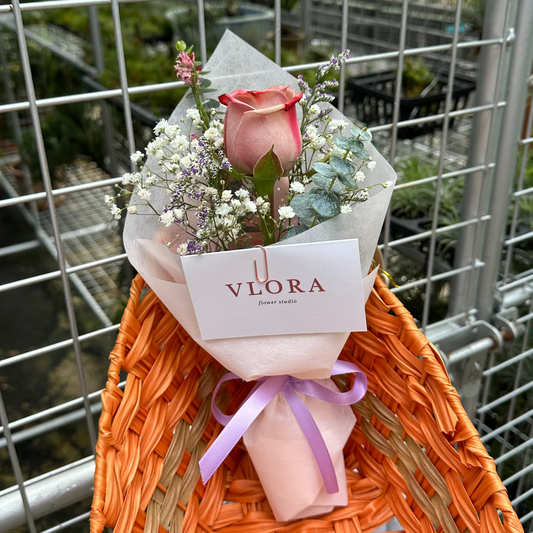 Small bouquet of flowers with a Cappuccino rose and baby's breath, wrapped in clear paper with a white card on an orange woven bag, against a metal fence background.