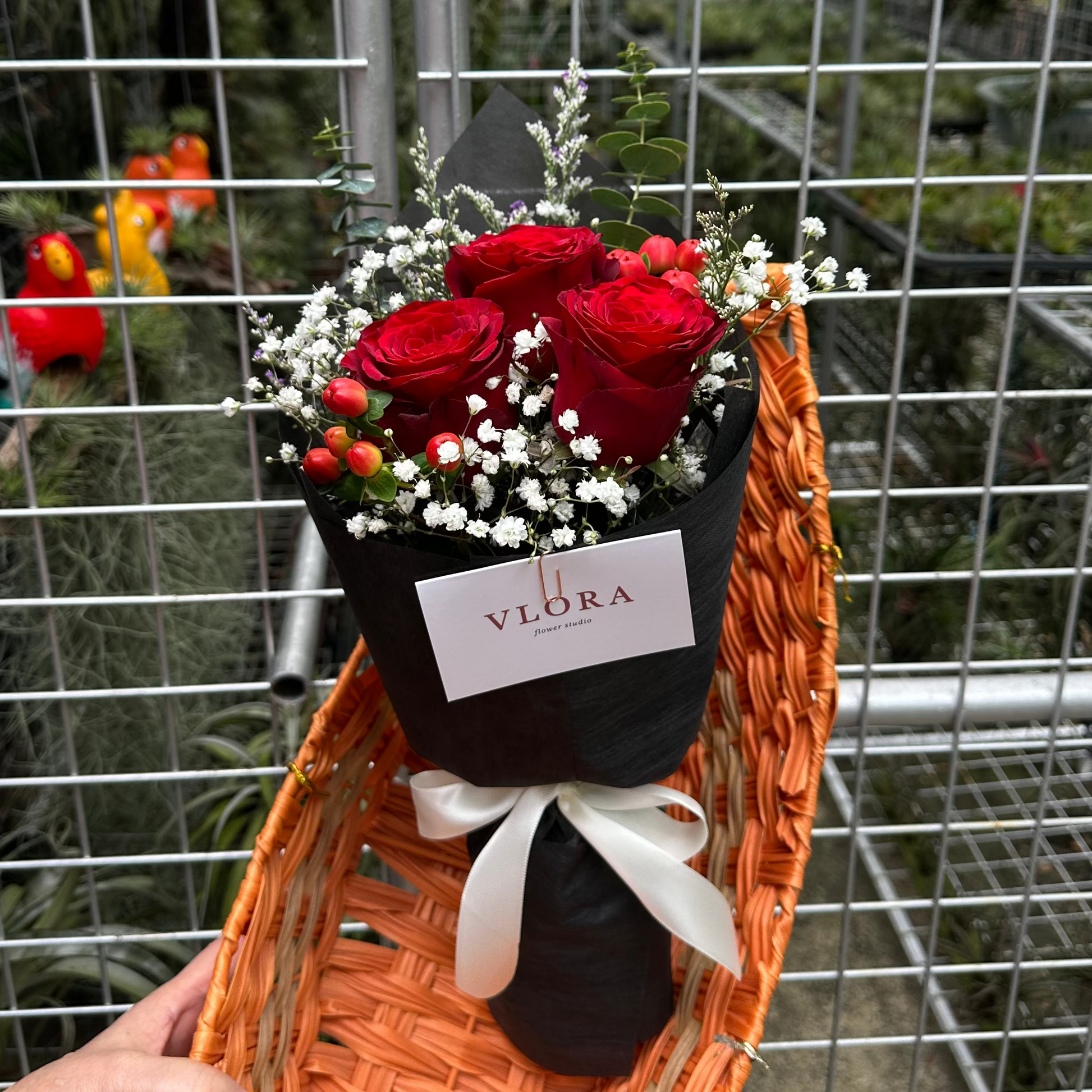 Bouquet of r3 ed roses in a black wrapping with 'VLORA' label, placed in a basket background.