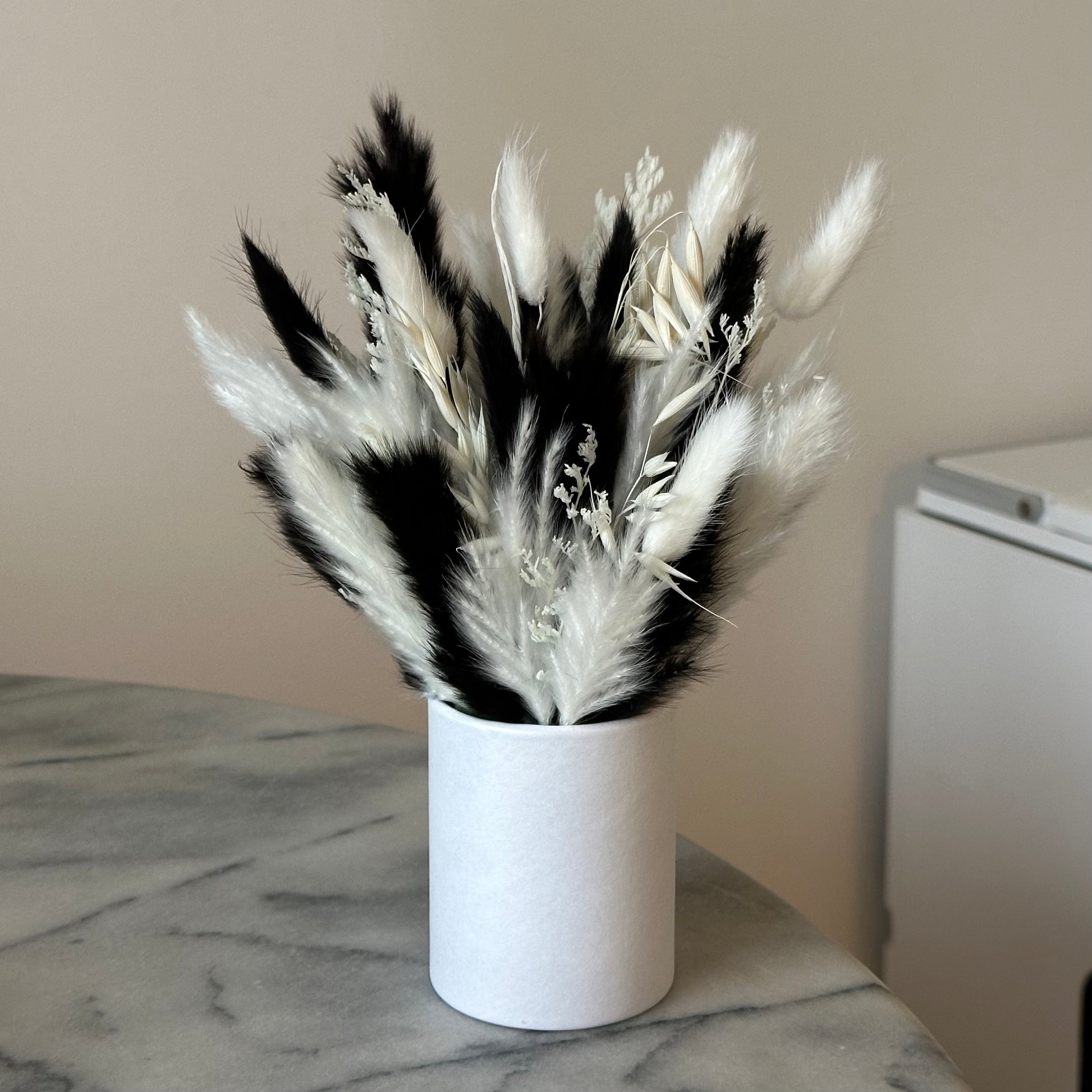 A preserved table arrangement featuring black and white Pampas and Bunny Tails in a white cylindrical vase, placed on a marble surface.