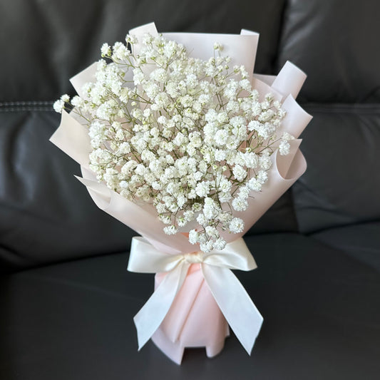 A bouquet of white Baby's Breath flowers wrapped with pink and white paper, presented on a dark couch.
