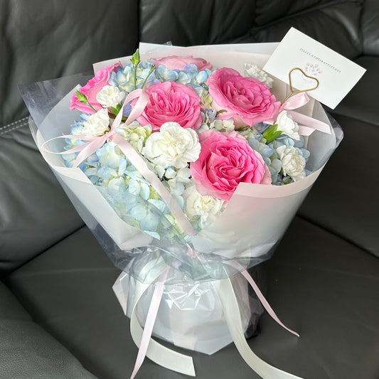 A fresh flower bouquet featuring pink roses, blue hydrangeas, white carnations, and decorative bows, wrapped in clear and pink paper.
