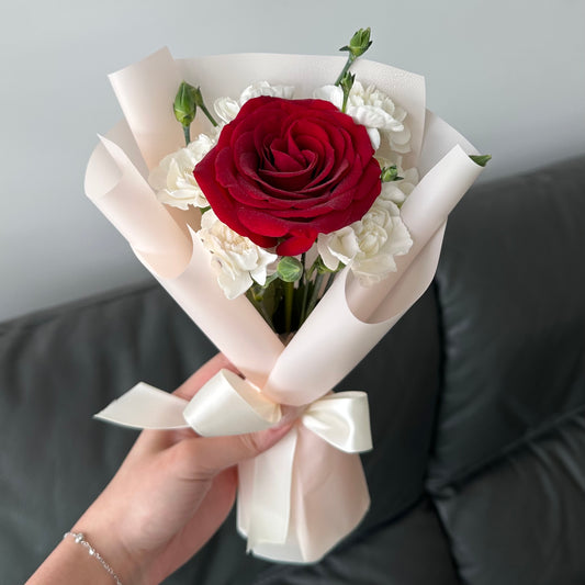A hand holding a bouquet with a single red rose and white carnations, wrapped in white paper with a ribbon.