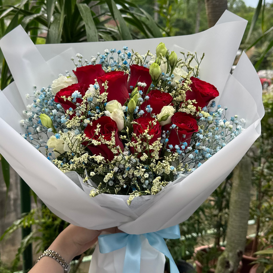 A hand holding a bouquet of red roses, baby's breath, and green foliage stems wrapped in white paper with a light blue ribbon, set against a background of potted plants.