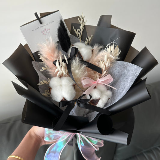 A preserved hand bouquet with white cotton flowers, fluffy bunny tails, and elegant pampas, wrapped in black with bows and a message card attached.