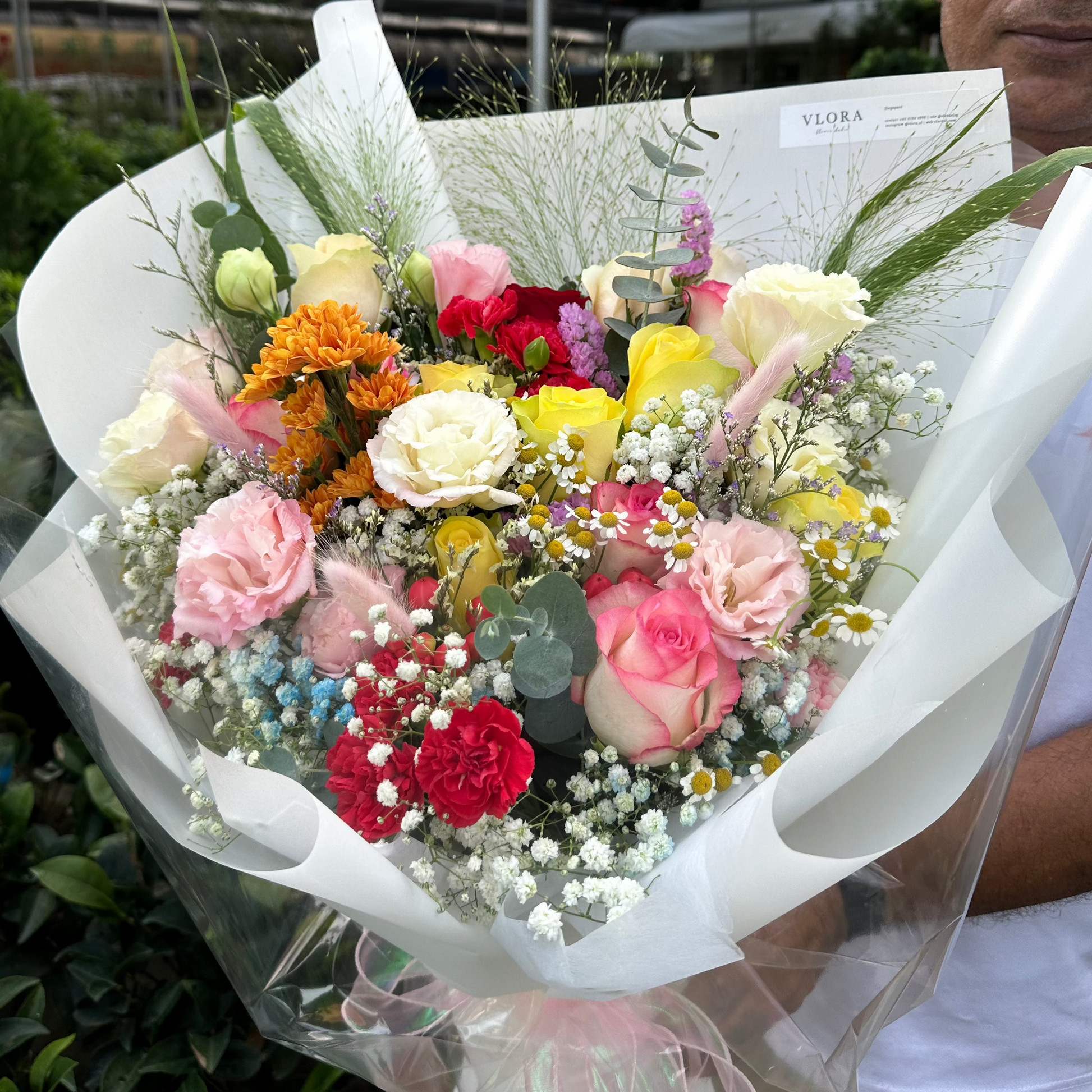 A vibrant mixed flower bouquet wrapped in white paper and clear cellophane, held by a man in a greenhouse setting.