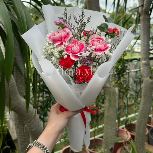 A hand holding a bouquet of pink carnations, red roses, and white baby's breath wrapped in white paper, set against a background of potted plants and trees.