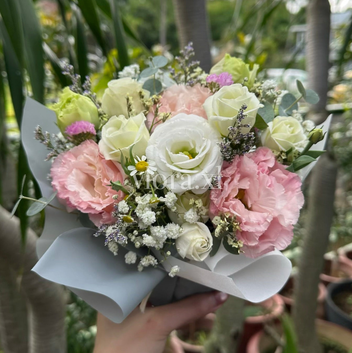 A hand holding a small bouquet of pink, white, and pale yellow flowers wrapped in light gray paper, photographed outdoors among potted plants and tree trunks.