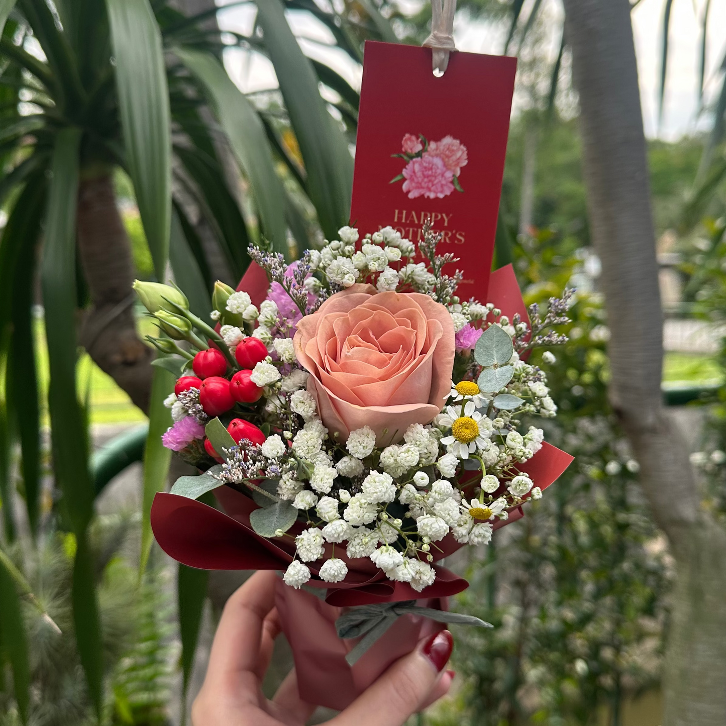 A hand holds a small floral bouquet featuring a peach rose, white baby's breath, red berries, and a red card in a garden setting with potted plants.
