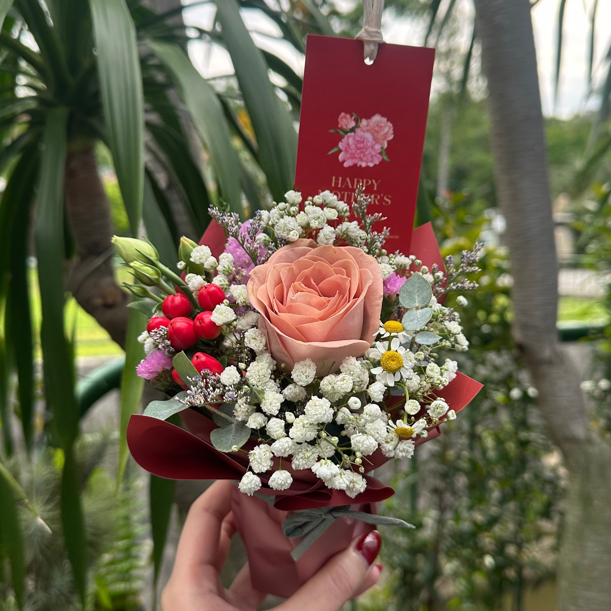 A hand holds a small floral bouquet featuring a peach rose, white baby's breath, red berries, and a red card in a garden setting with potted plants.