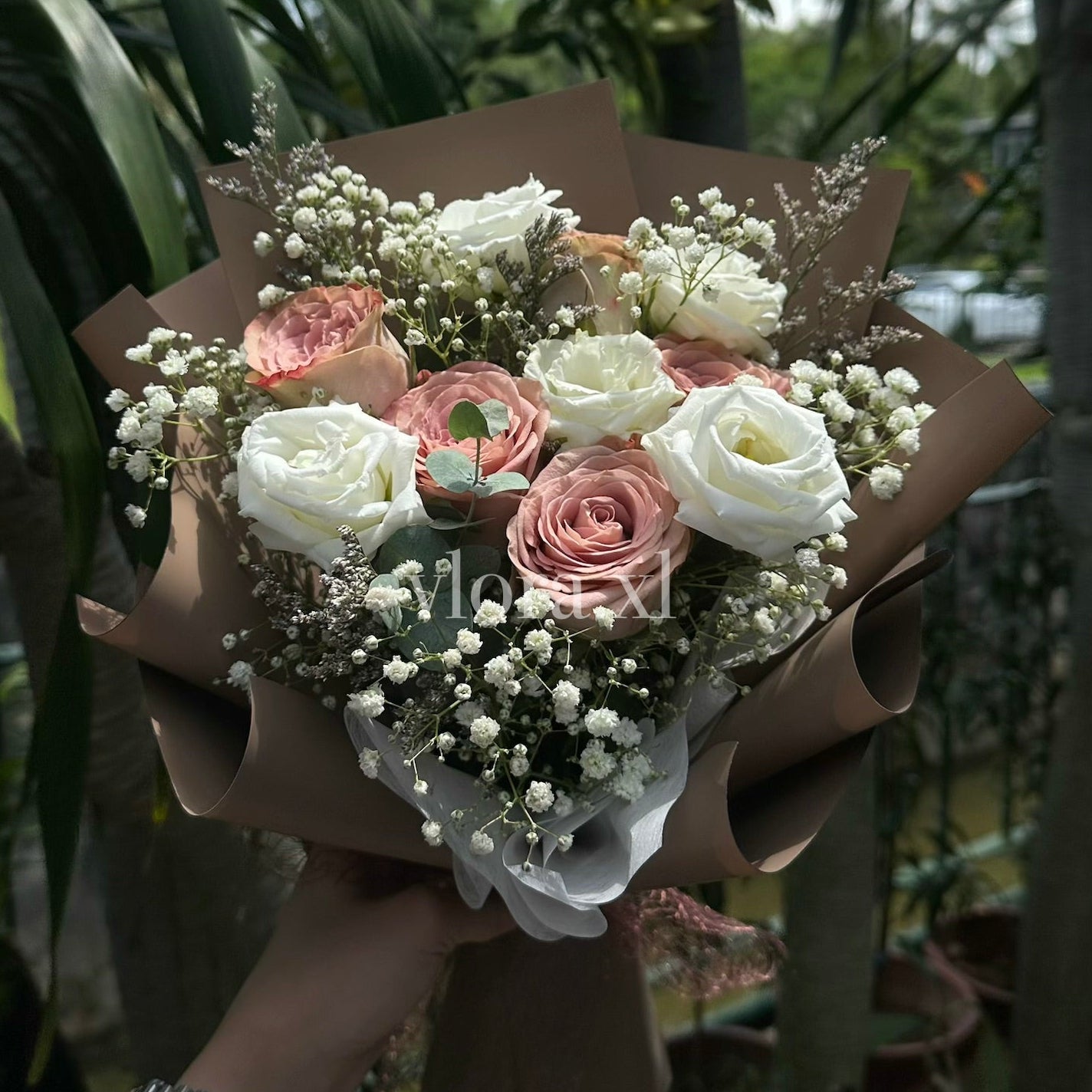 A hand holding a bouquet of pink and white roses with baby's breath flowers, wrapped in brown paper, with a real garden background.