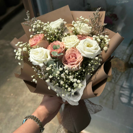 A hand holding a bouquet of artificial flowers, including white and pink roses, baby's breath, and green foliage, wrapped in brown paper.