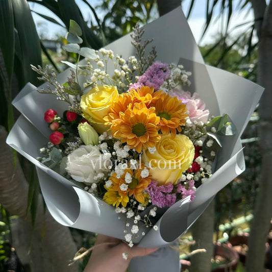 A hand holding a wrapped flower bouquet outdoors, featuring yellow roses, orange daisies, white carnations, purple filler flowers, and small red berries in gray paper wrapping.