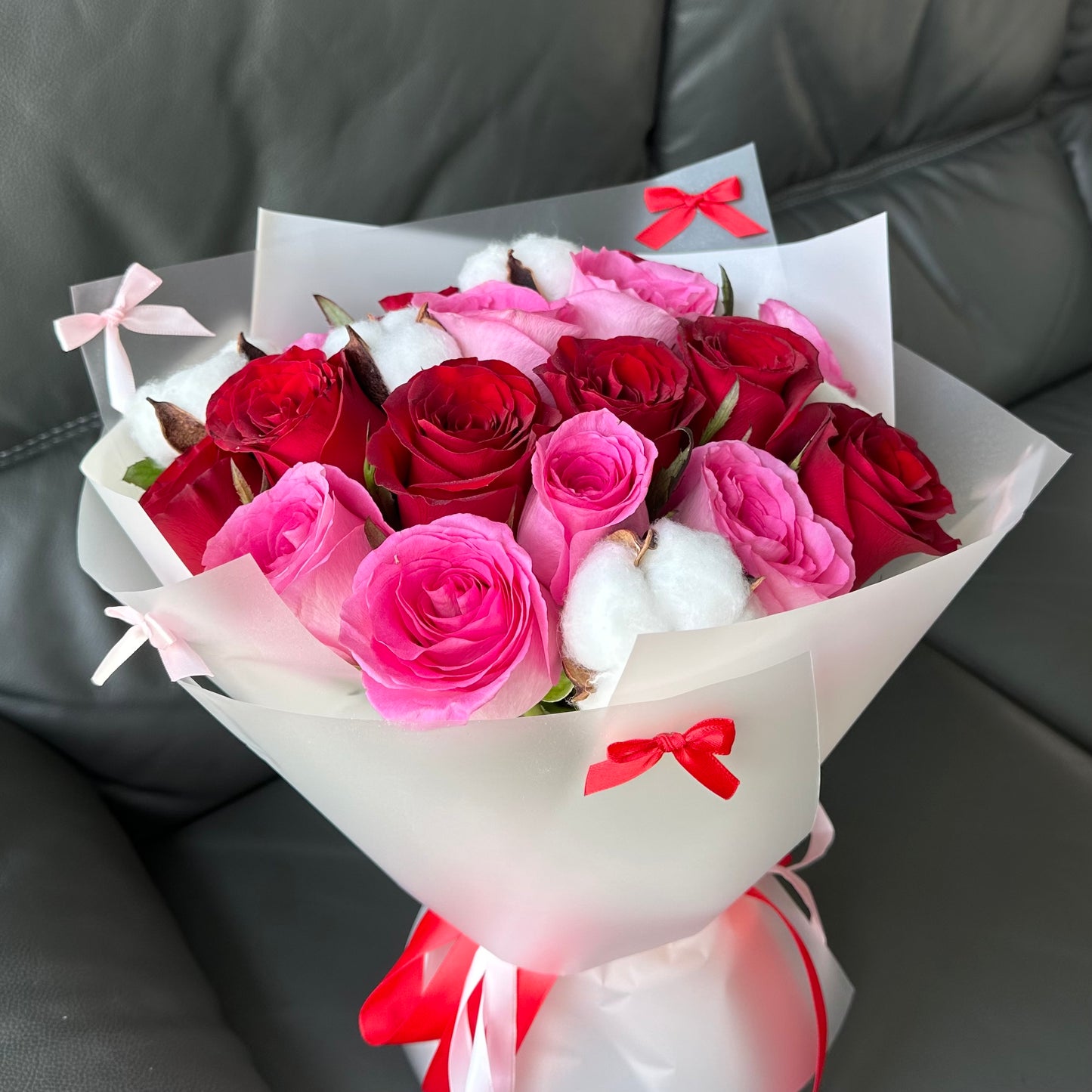 A bouquet of flowers consisting of red, pink, and white roses, wrapped in white paper with pink and red decorative bows.
