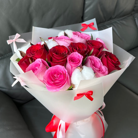 A bouquet of flowers consisting of red, pink, and white roses, wrapped in white paper with pink and red decorative bows.