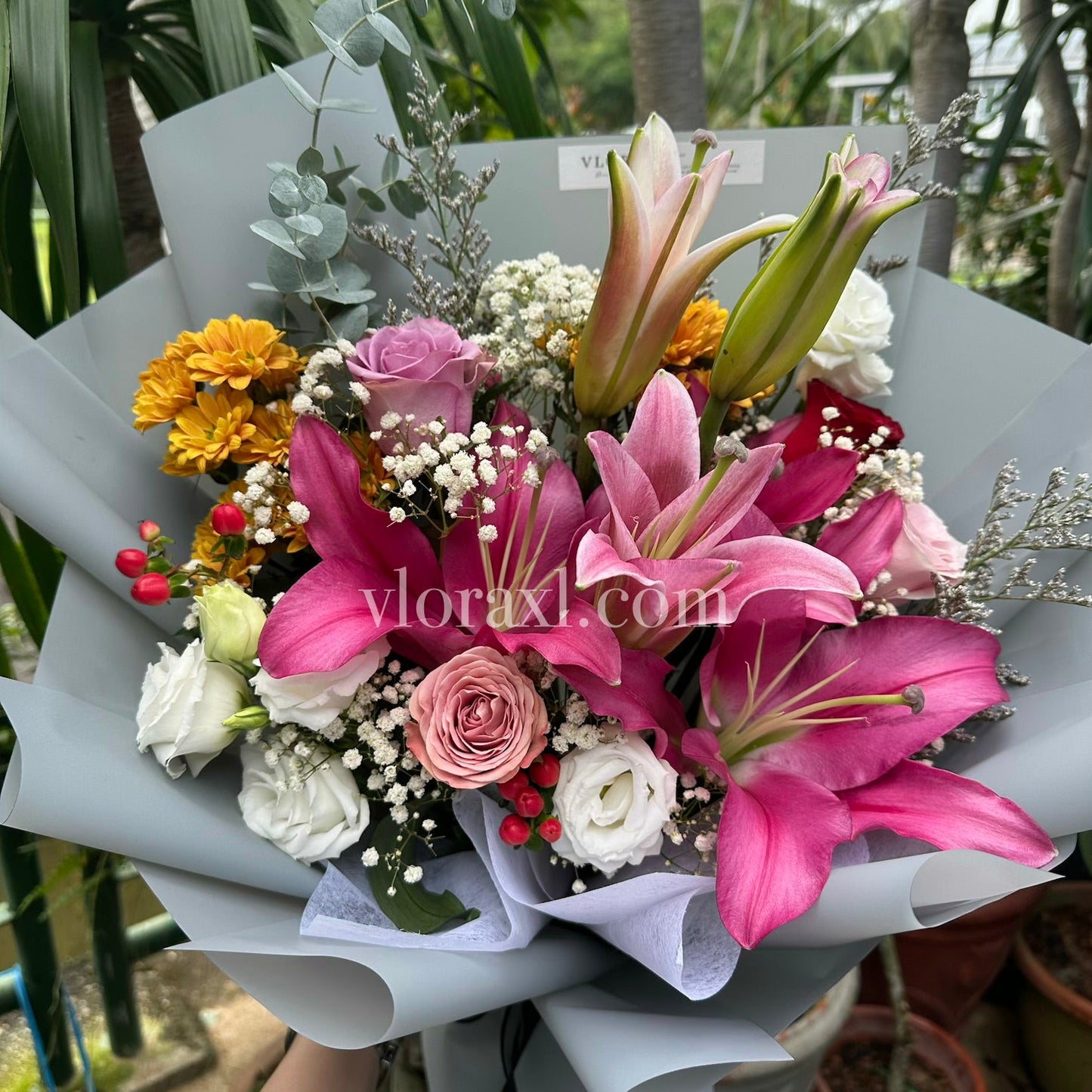 A hand-held flower bouquet featuring vibrant pink lilies, pink and white roses, yellow marigolds, and white baby's breath, wrapped in light gray paper, with a garden background.