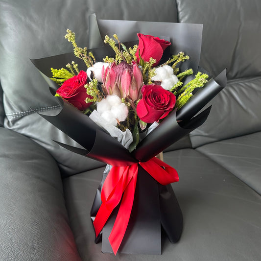 A bouquet of flowers including red roses, a protea, and white cotton flowers, wrapped in black paper with a red ribbon, displayed on a couch.