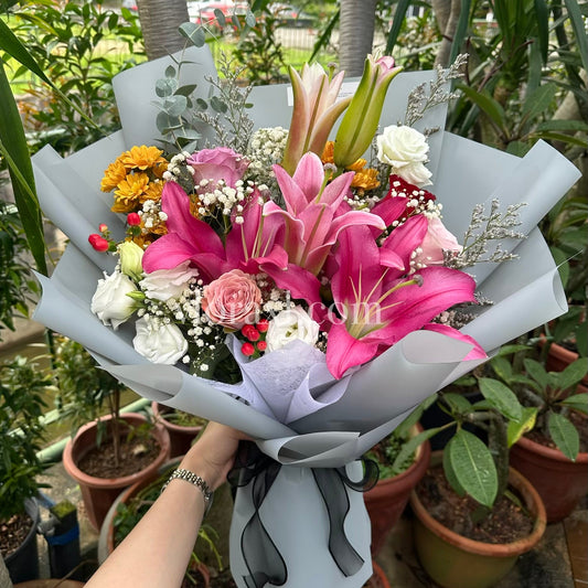A hand holding a vibrant flower bouquet in a garden setting. The bouquet features bright pink lilies, pink roses, white carnations, yellow daisies, and green foliage, wrapped in light gray paper with a black ribbon.