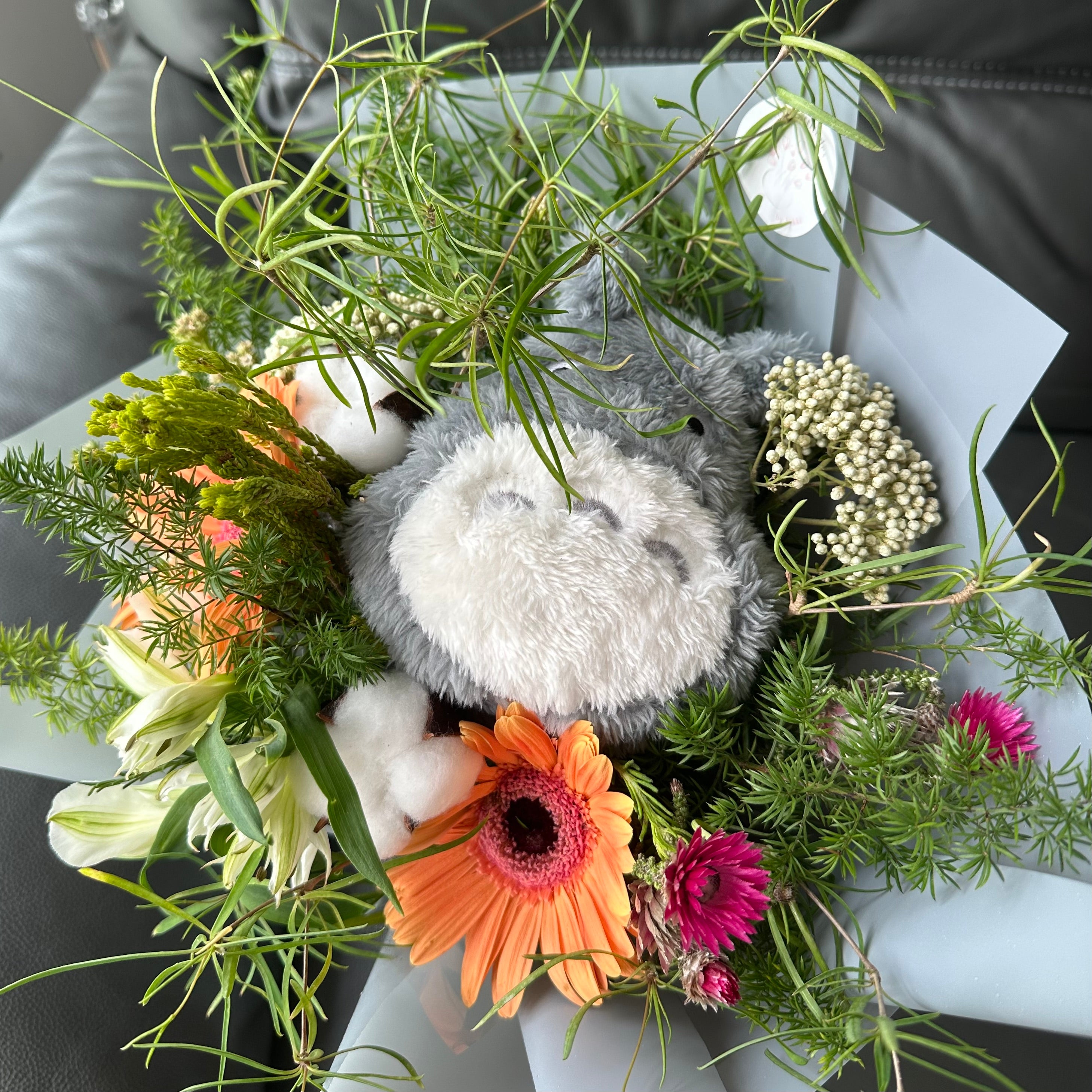 A bouquet of flowers with a Totoro plush toy character included, wrapped in grey.