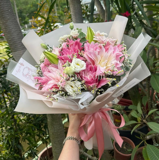 A hand holding a bouquet of pink and white flowers wrapped in white paper with a pink ribbon, set against a background of potted plants and trees.