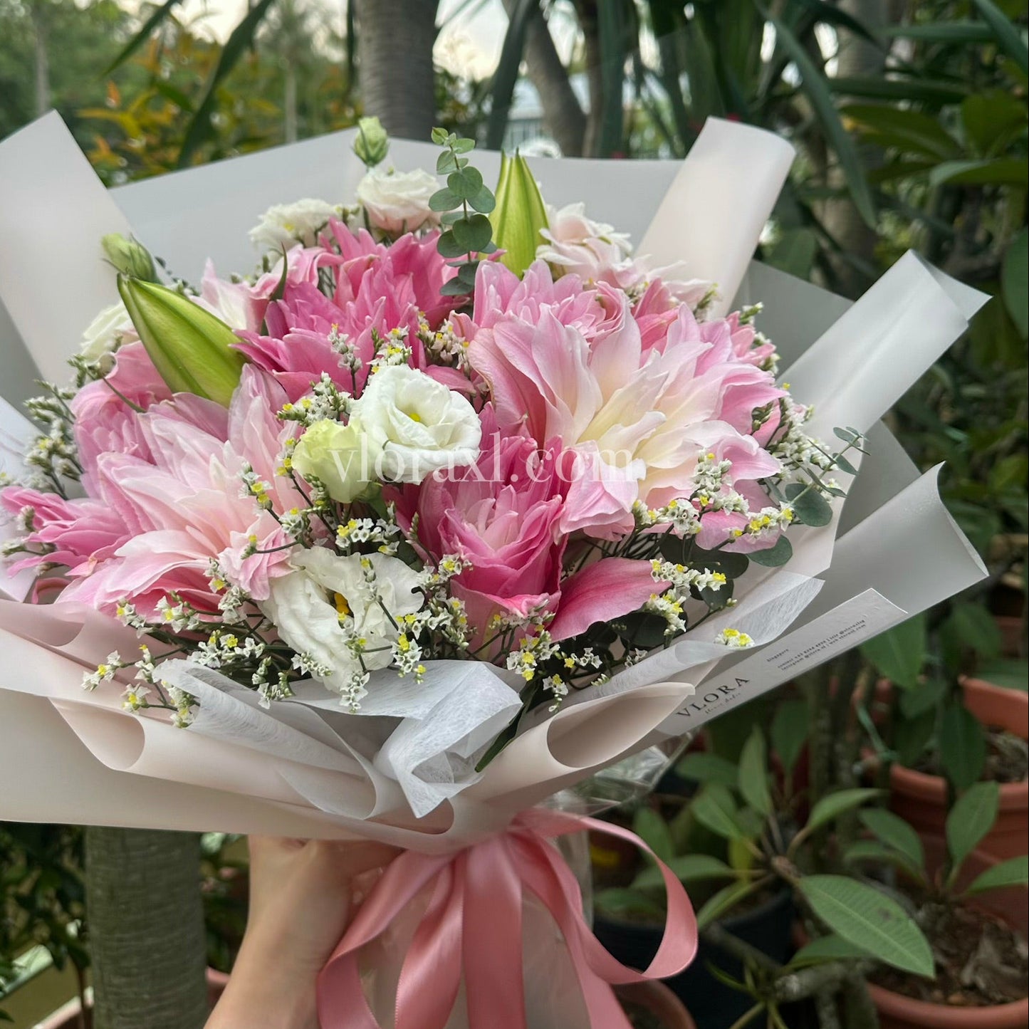 A hand-held bouquet of pink and white flowers wrapped in white paper with a pink ribbon, photographed outdoors among green potted plants.