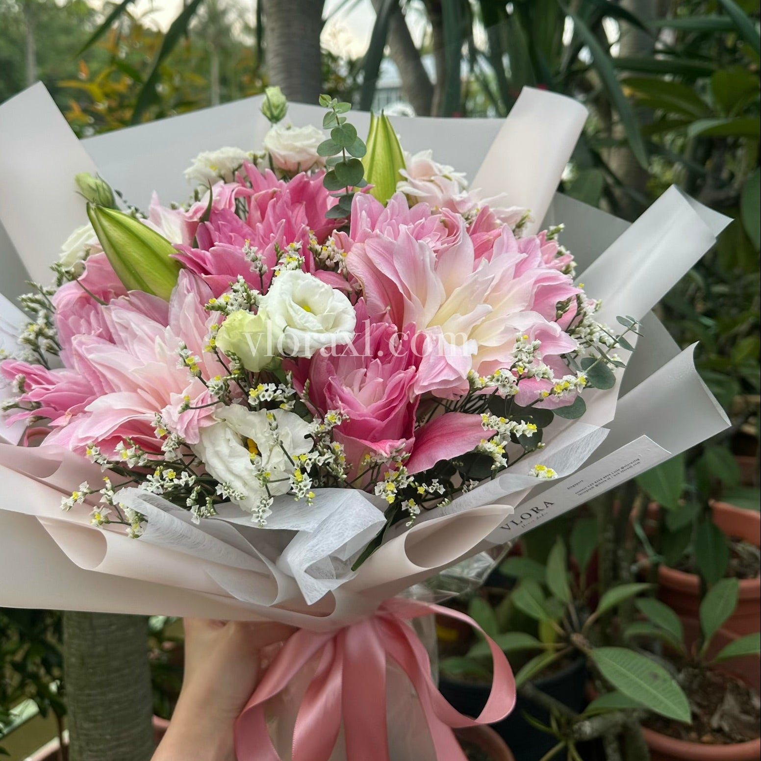 A hand-held bouquet of pink and white flowers wrapped in white paper with a pink ribbon, photographed outdoors among green potted plants.