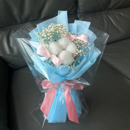 A bouquet of cotton flowers and baby's breath arranged with blue and pink bows, placed on a sofa.