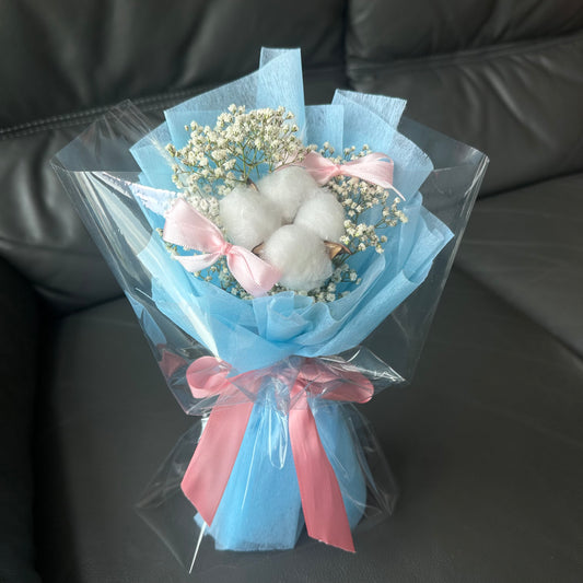 A bouquet of cotton flowers and baby's breath arranged with blue and pink bows, placed on a sofa.