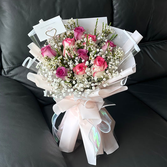 A bouquet of flowers consisting of soft pink Roses and elegant purple Roses, wrapped in light pink paper with a ribbon, placed on a black surface.