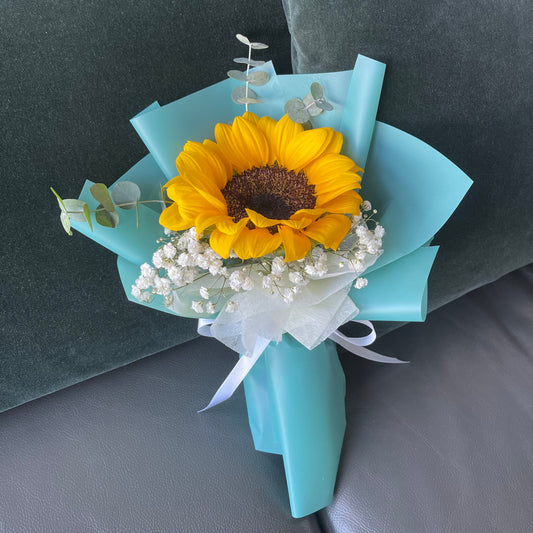 A bouquet featuring a yellow sunflower and Baby’s Breath, wrapped in turquoise paper with a white ribbon.
