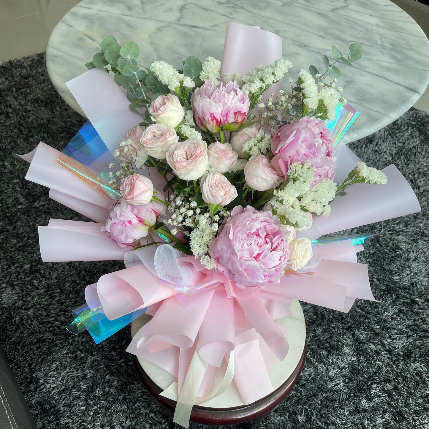 A floral arrangement featuring pink peonies and roses with light pink and iridescent wrapping.