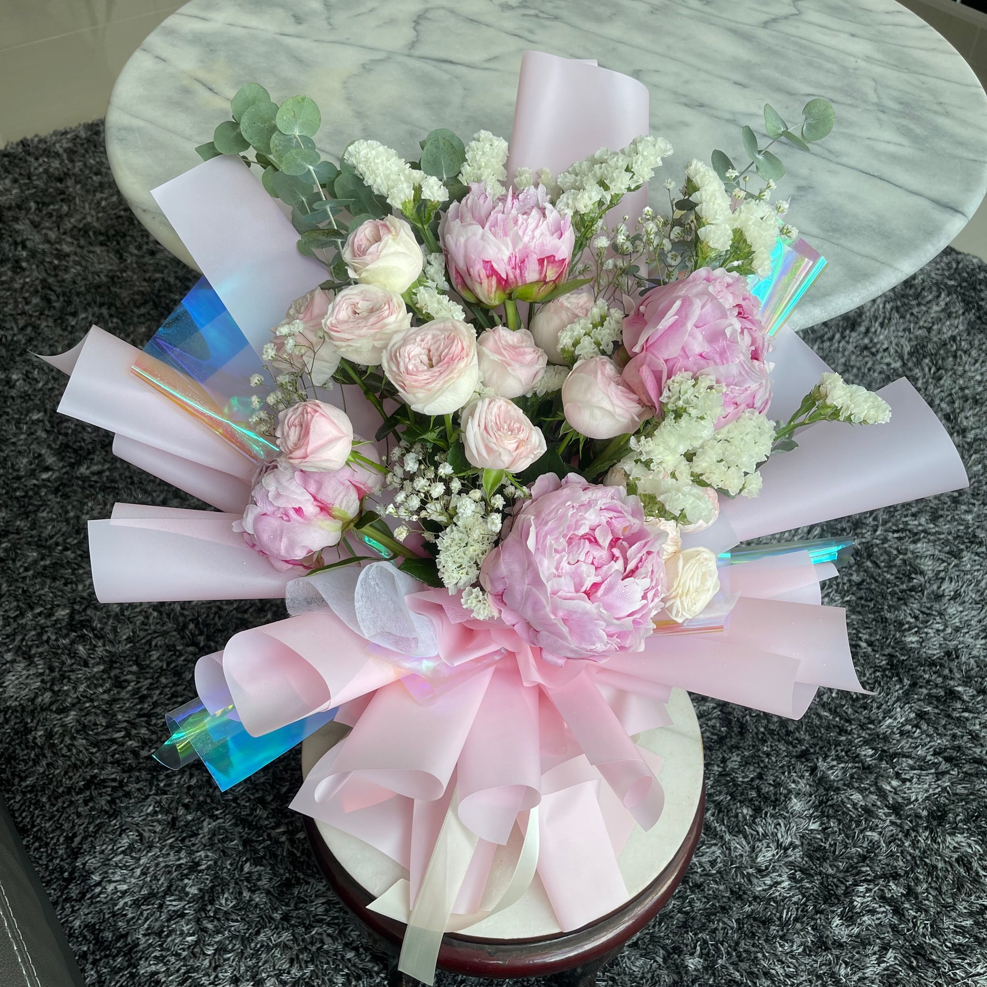 A floral arrangement featuring pink peonies and roses with light pink and iridescent wrapping.