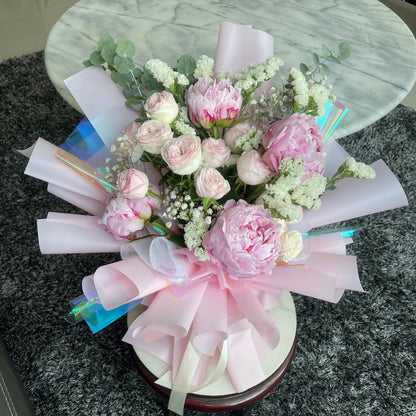 A floral arrangement featuring pink peonies and roses with light pink and iridescent wrapping.